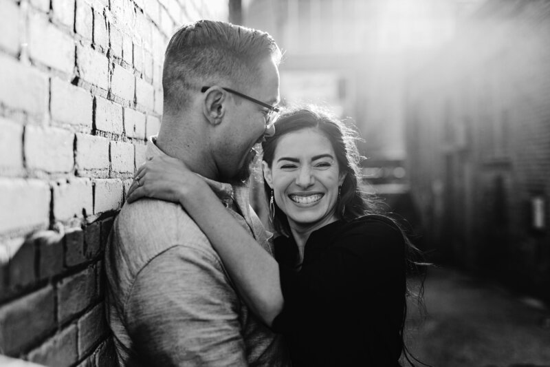 Black-and-white portrait of a couple embracing in an alleyway, with the woman smiling toward the camera.