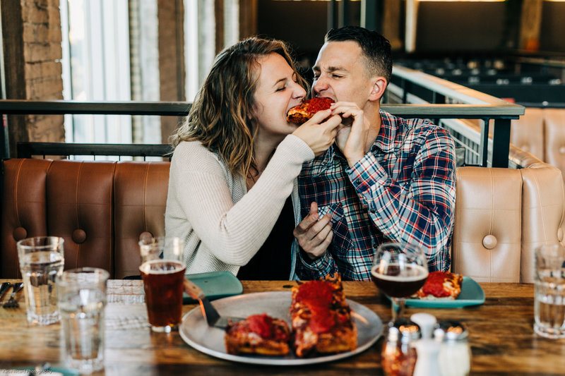 Couple sharing a slice of pizza at a restaurant booth, with drinks and deep-dish pizza on the table.