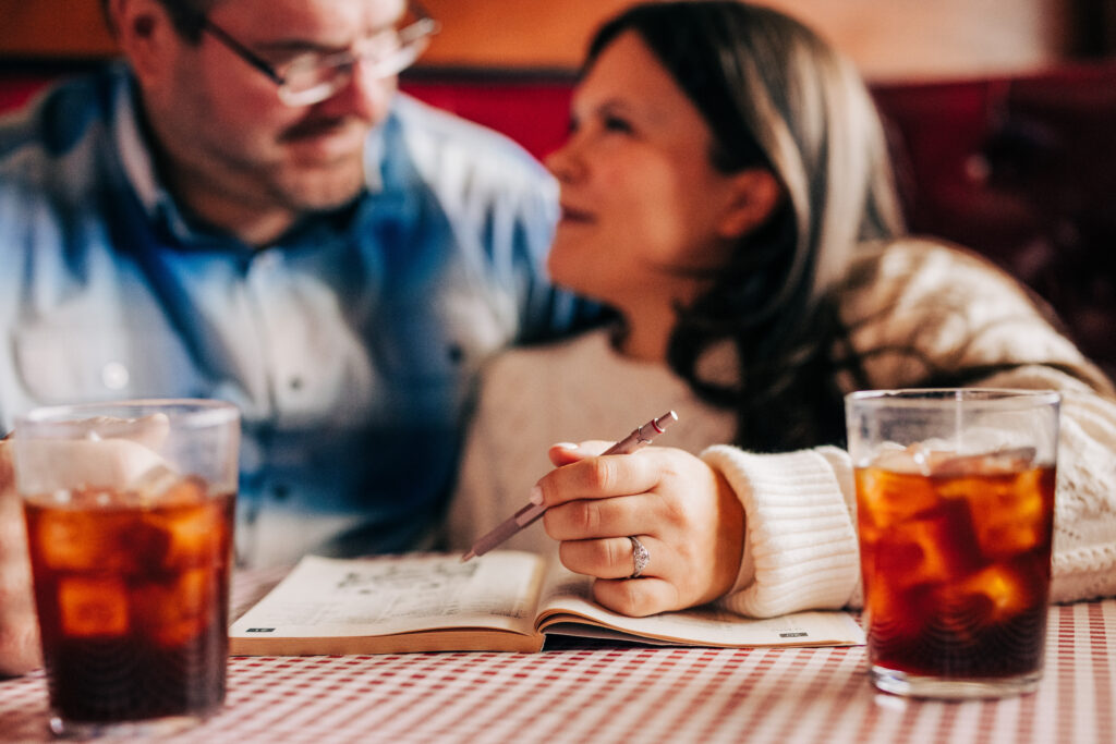 Couple seated at a restaurant booth looking at a notebook together, with the woman holding a pen and iced drinks on the table.