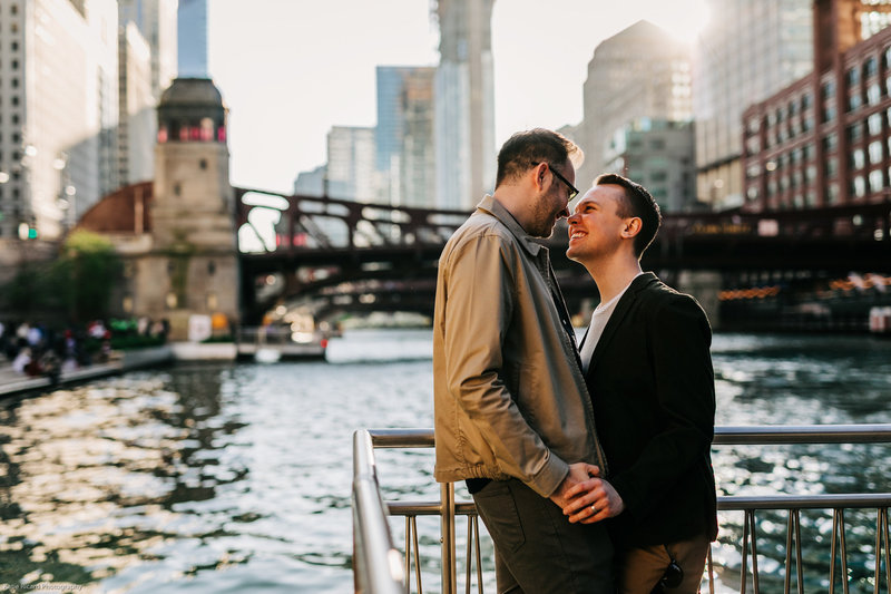 Couple holding hands along the Chicago Riverwalk at sunset, with city skyscrapers and a bridge in the background.