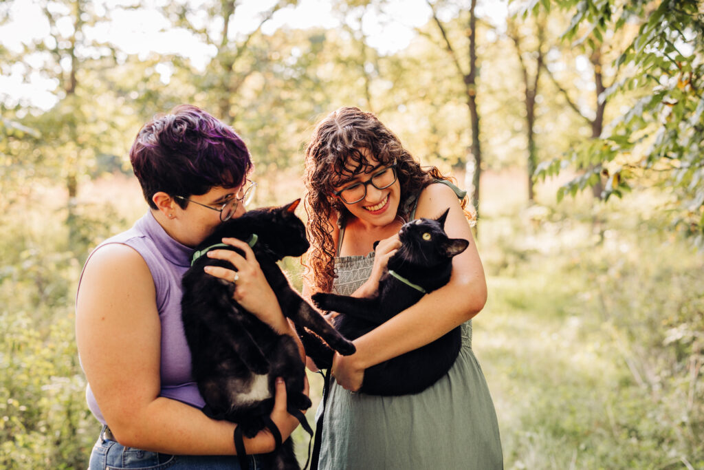 Two people smiling at each other while holding black cats in a wooded, sunlit outdoor setting.