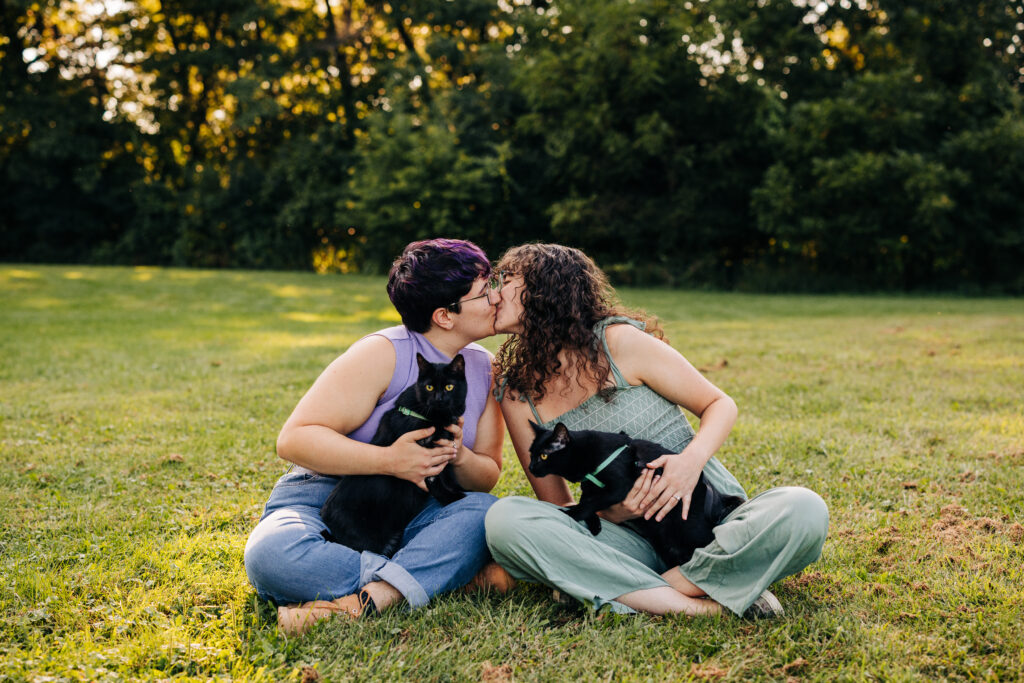 Two people sit cross-legged in an open grassy area, sharing a kiss while holding their black cats.