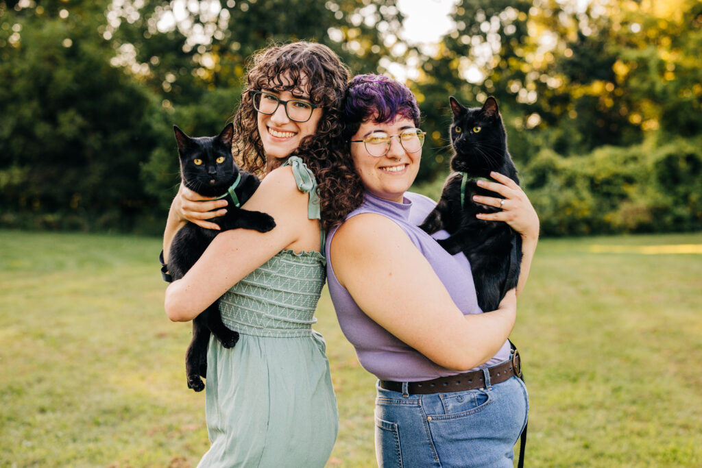 Two people pose back-to-back in a field, each smiling and holding a black cat in their arms.