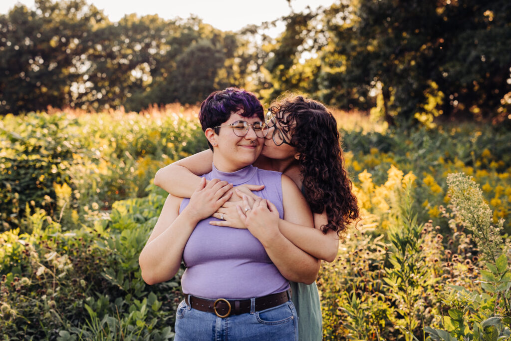 A person wraps their arms around their partner from behind and kisses their cheek in a sunlit field with tall grasses.