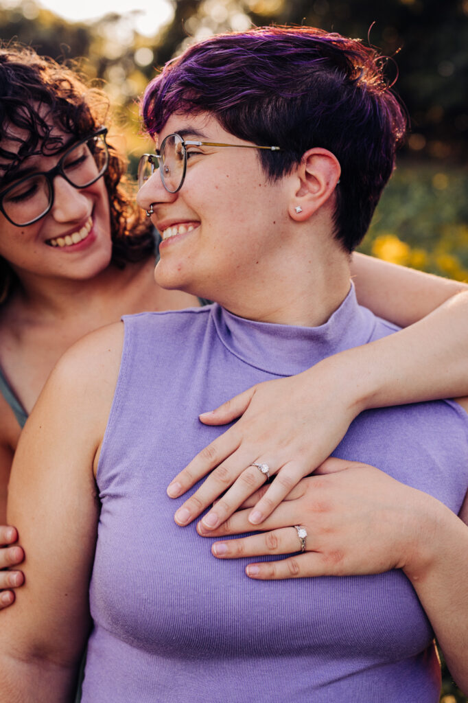 Tight crop of two people smiling closely with hands over each other’s hearts, showing matching engagement rings.