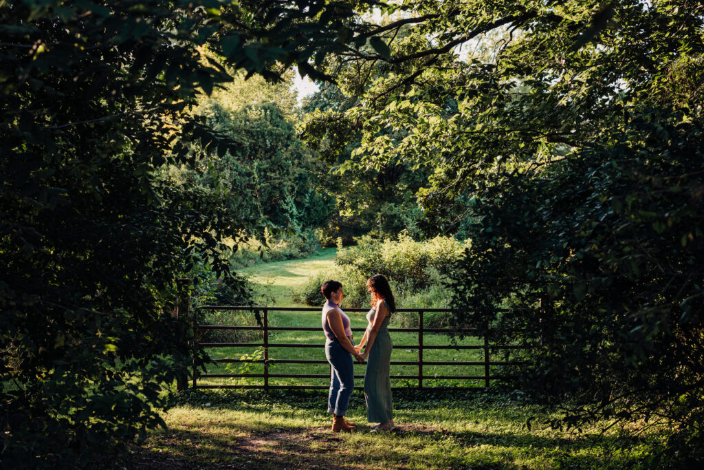 Couple stands hand-in-hand under leafy trees near a rustic gate, framed by sunlight filtering through the branches.