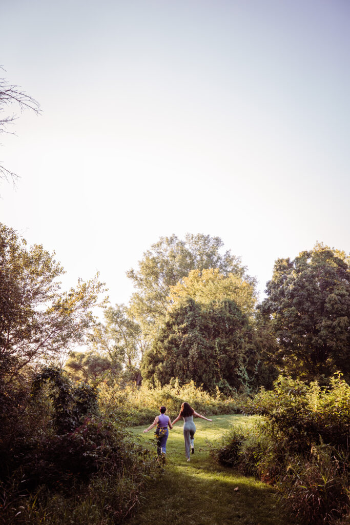 Two people holding hands, running away down a grassy path surrounded by dense greenery and tall trees. The sunlight filters softly through the trees as they head into the distance, arms gently outstretched.