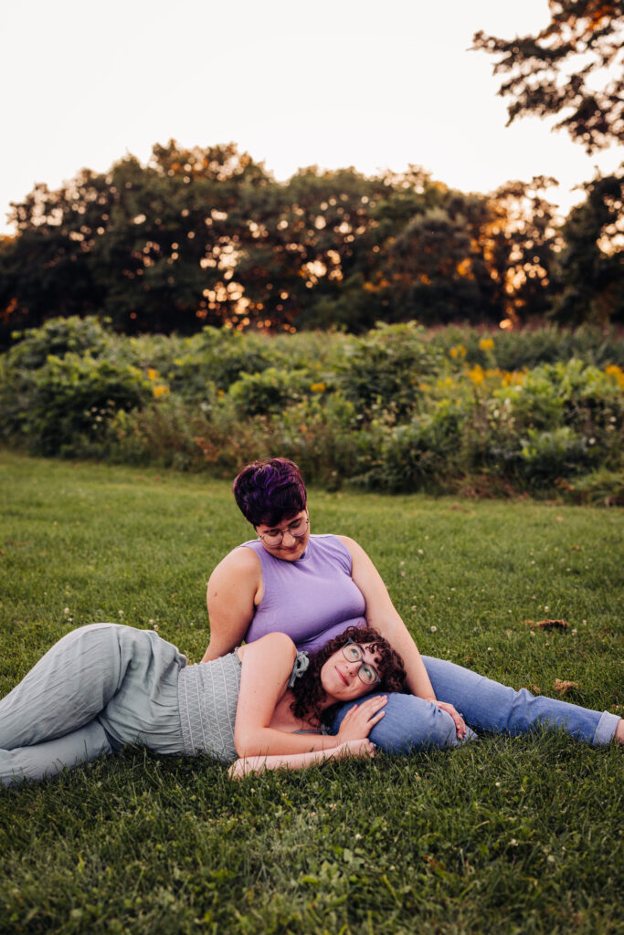 One person sits on the grass while the other lies with their head in their partner's lap, gazing softly at the camera. The background glows with wildflowers and trees lit by a warm sunset.