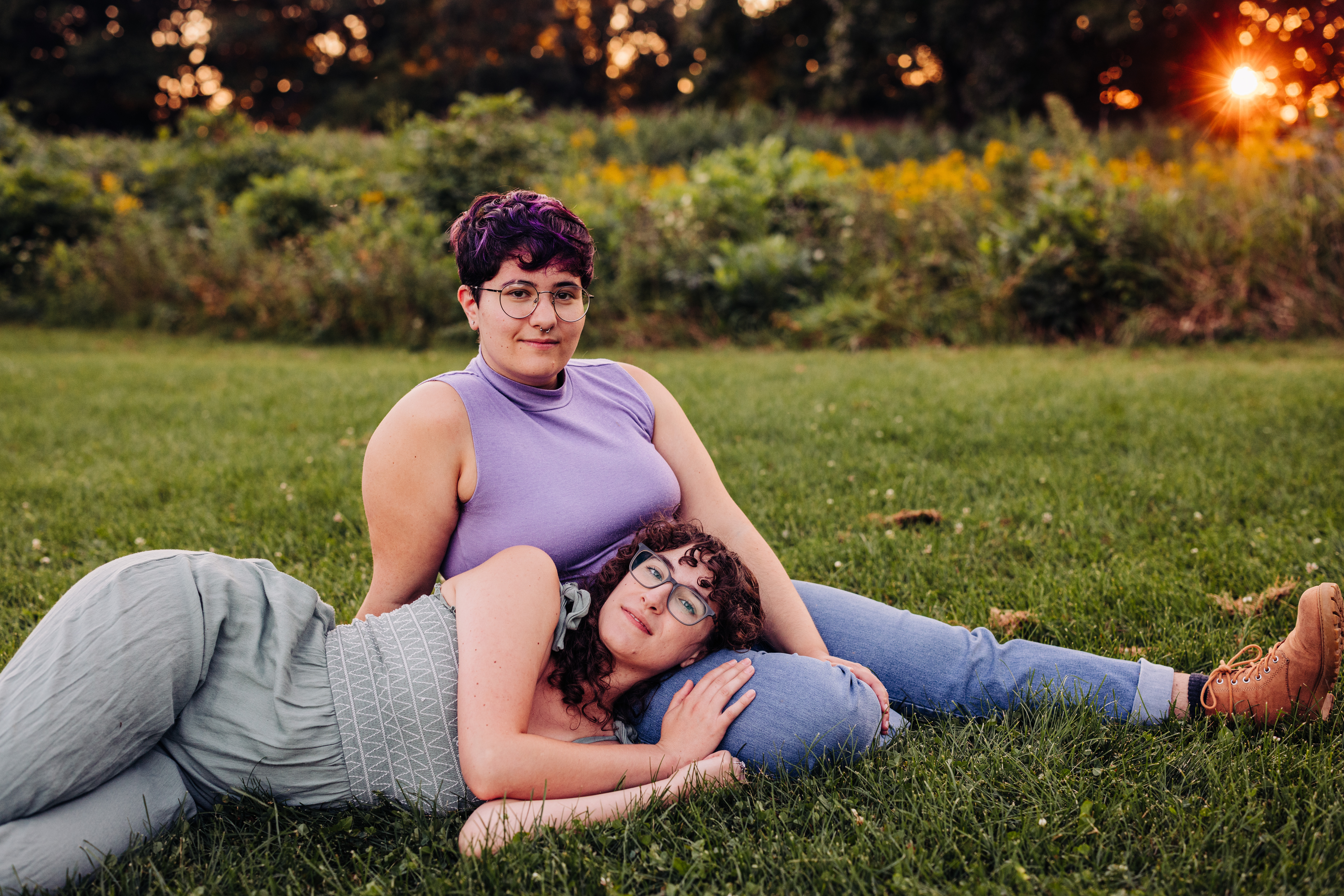 Two people sit on the grass during golden hour. One reclines with her head on the other’s lap, looking directly at the camera, while her partner sits upright beside her, also facing forward with a calm expression.