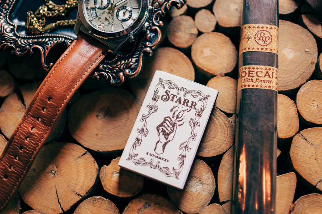 Groom details including a leather watch, cigar, and custom matchbox styled on stacked wood slices.