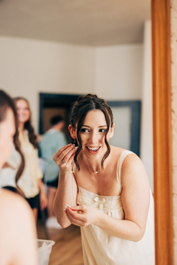 Bride smiling while putting on earrings during wedding morning preparations in a softly lit room.