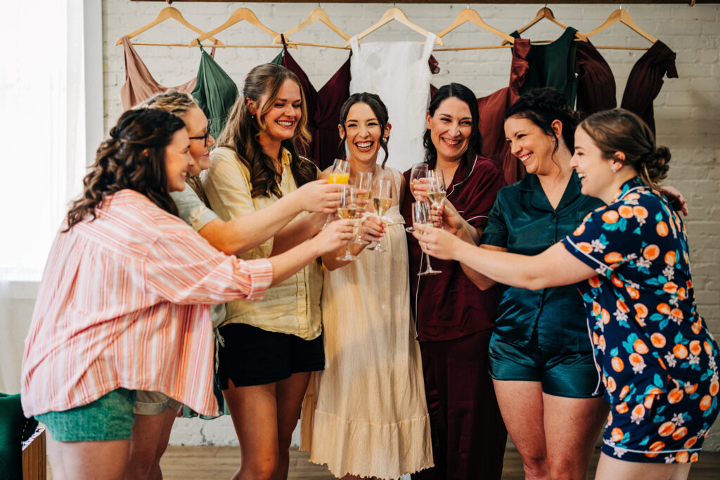 Bridal party clinking champagne glasses while getting ready, with bridesmaid dresses hanging in the background.