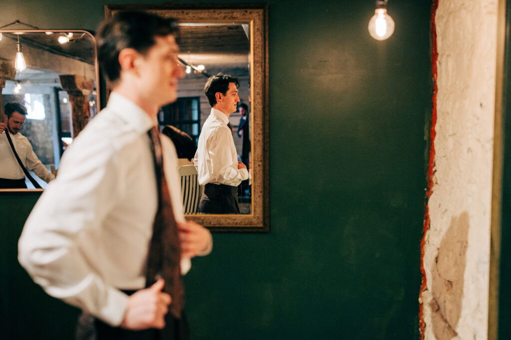 Groom seen adjusting his tie reflected in a mirror against green walls and exposed brick.