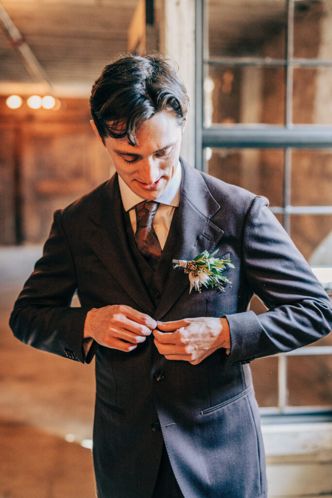 Groom buttoning his suit jacket indoors, wearing a boutonniere with greenery and neutral florals.