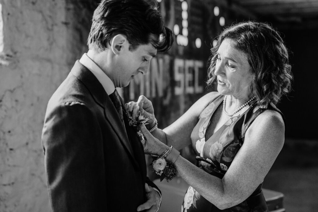 Black and white photo of groom’s mother pinning his boutonniere during wedding preparations at The Lageret.