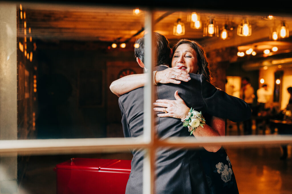 Mother hugging her son before the ceremony, photographed through a window inside a warmly lit wedding venue.