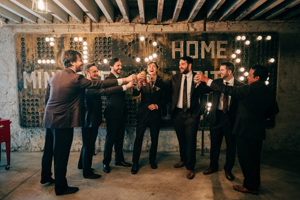 Groom and groomsmen laughing and raising drinks together in front of a vintage sign inside The Lageret.