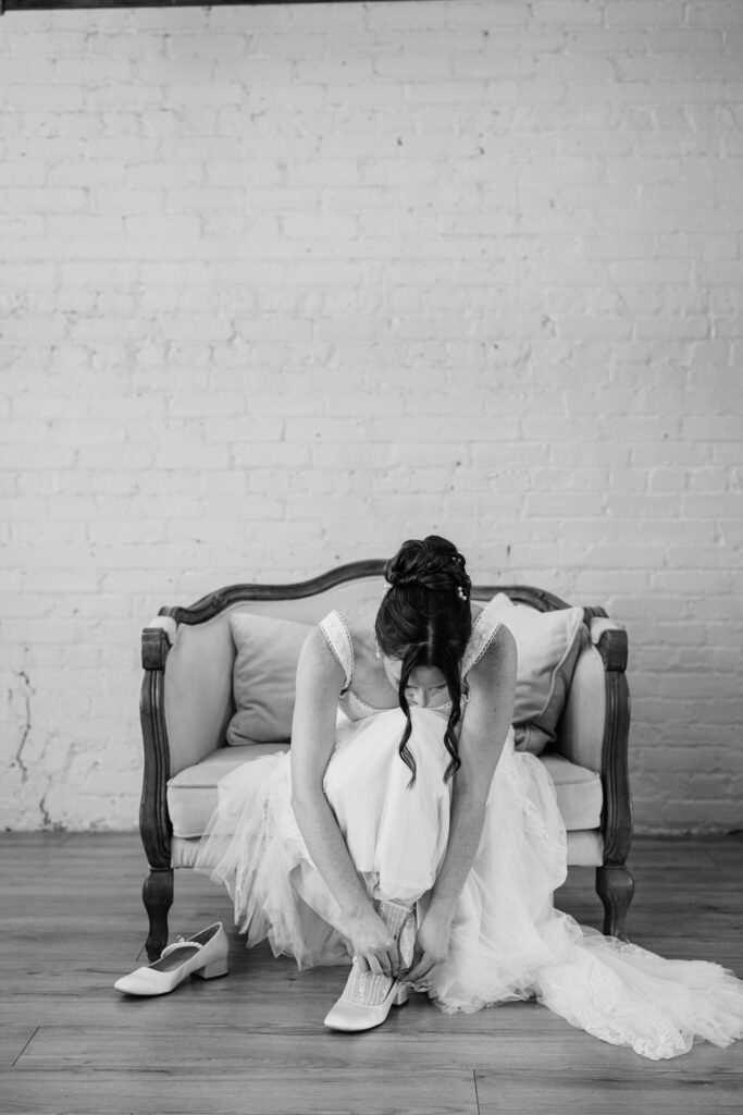Black and white photo of bride sitting on a vintage couch while fastening her wedding shoes.
