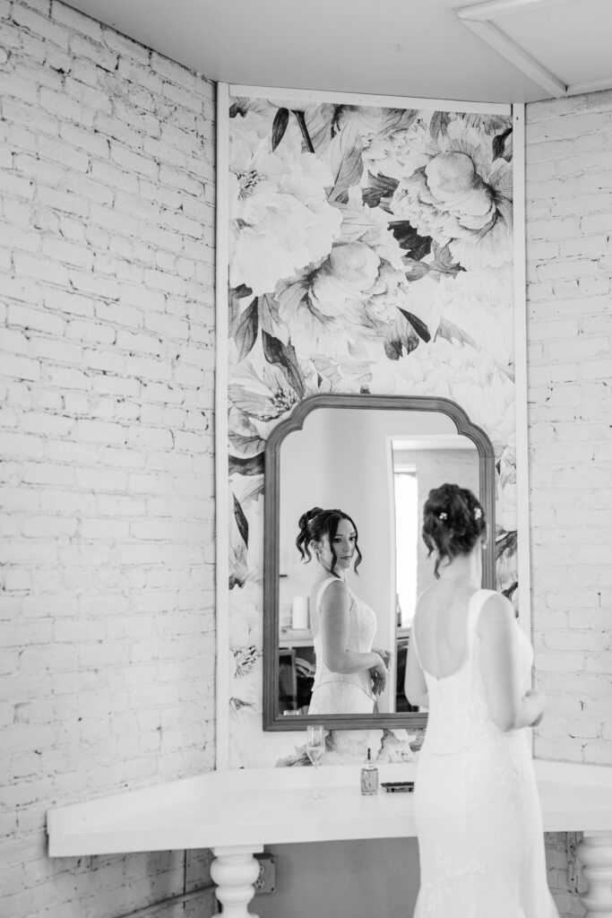Bride standing at a vanity and looking at her reflection in a mirror, surrounded by textured brick walls.