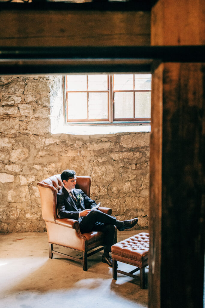 Groom sitting in a leather chair reading a letter during a quiet moment before the ceremony.