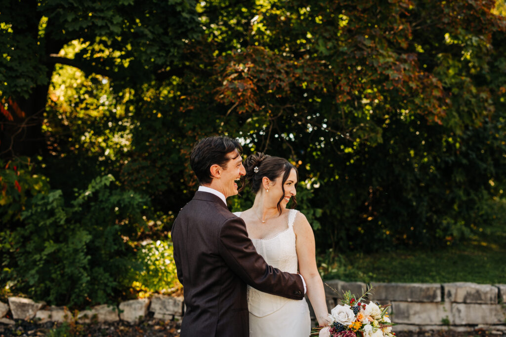Bride and groom laughing side by side after their first look, framed by trees at a fall Wisconsin wedding.