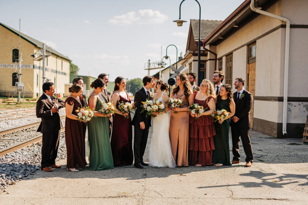 Bride and groom kissing while surrounded by their wedding party near historic train tracks in Stoughton, Wisconsin.