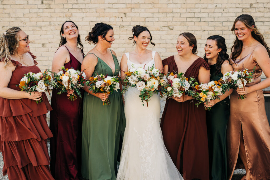 Bridesmaids laughing together while holding bouquets against a brick wall at an outdoor Wisconsin wedding.