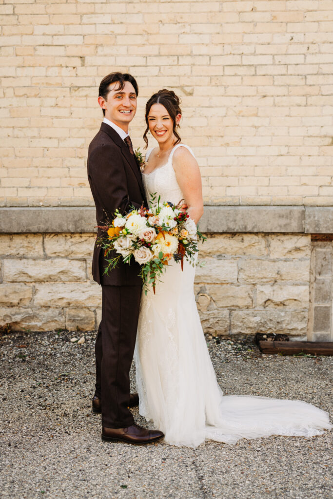 Bride and groom smiling together during wedding portraits against a cream brick wall at The Lageret in Wisconsin.