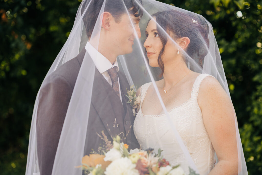 Bride and groom standing close under the veil during romantic outdoor wedding portraits surrounded by greenery.