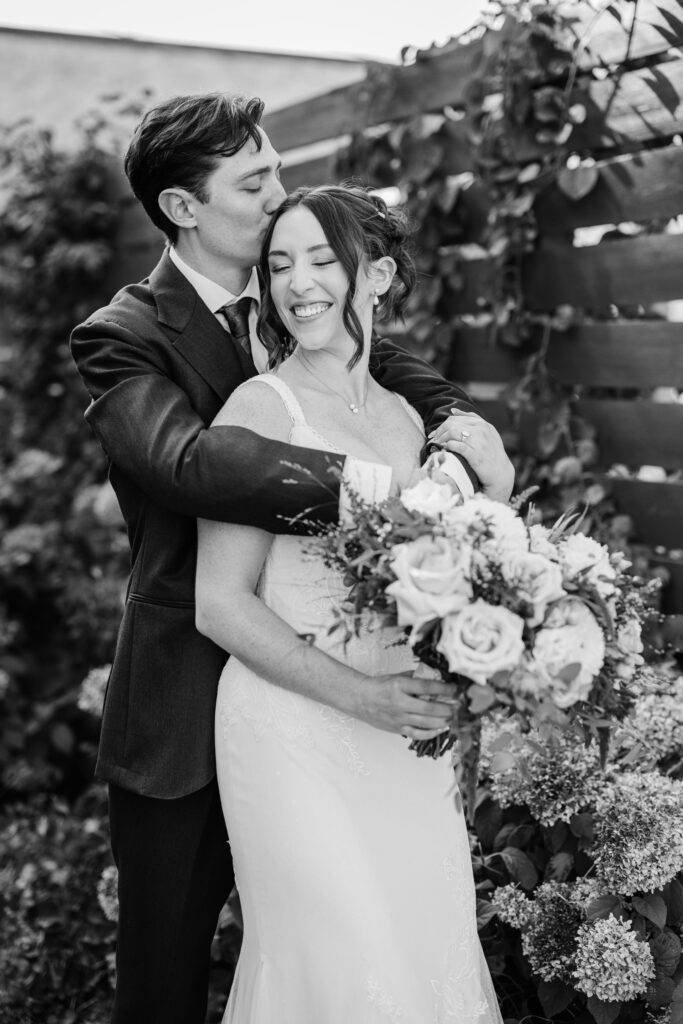 Black and white portrait of groom hugging the bride from behind while she smiles, holding her bouquet outdoors.