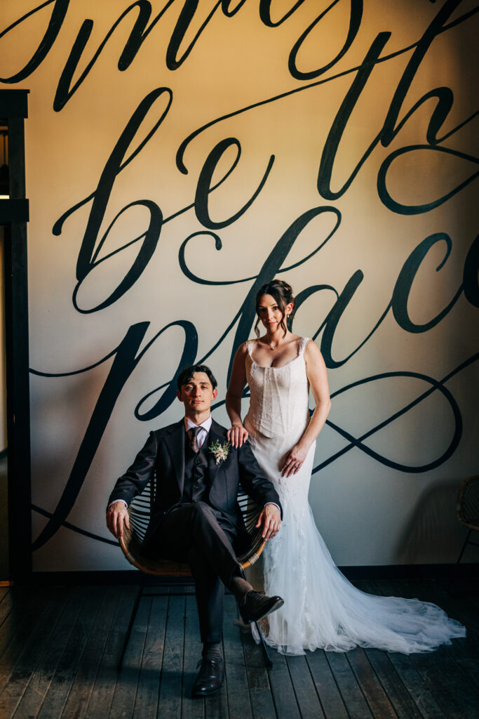 Bride standing beside seated groom during indoor wedding portraits in front of a painted mural wall at The Lageret.