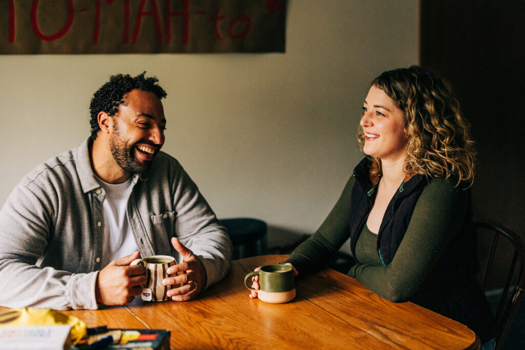 Two adults sitting at a wooden table indoors, smiling and talking while holding mugs of coffee.