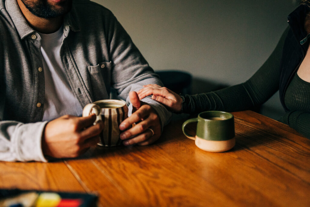 Close-up of two people’s hands at a wooden table, one hand resting on the other beside coffee mugs.