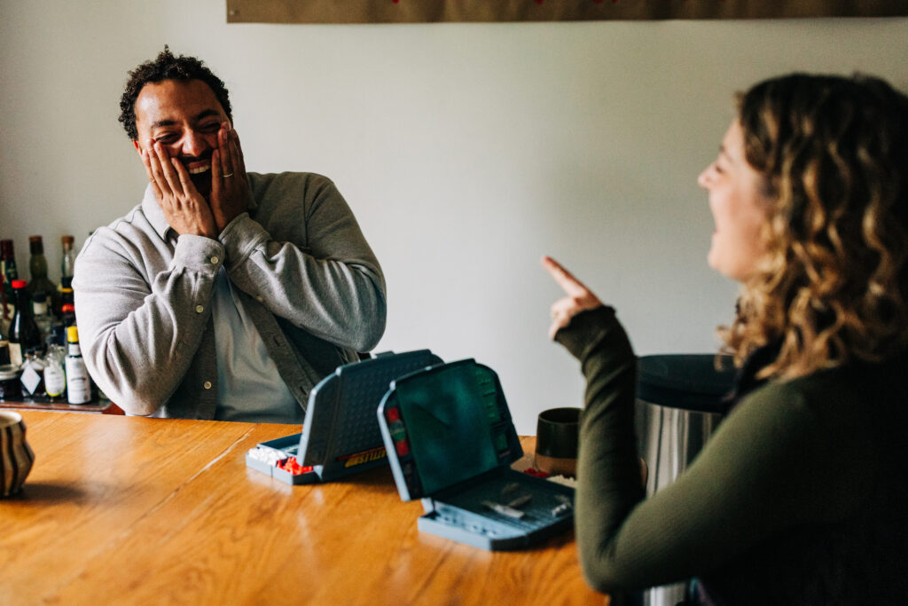 Man laughing at a table while holding a coffee mug, with a board game open in front of him.