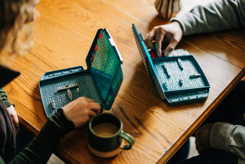 Overhead view of two people playing a board game on a wooden table with coffee mugs nearby.