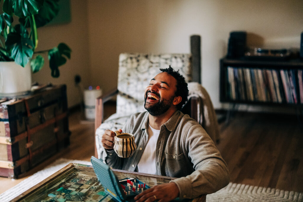 Man sitting in a living room laughing while holding a coffee mug, with a board game on the table.
