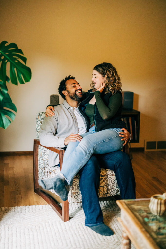 Couple sitting together in a patterned armchair at home, smiling and leaning into each other in soft window light.