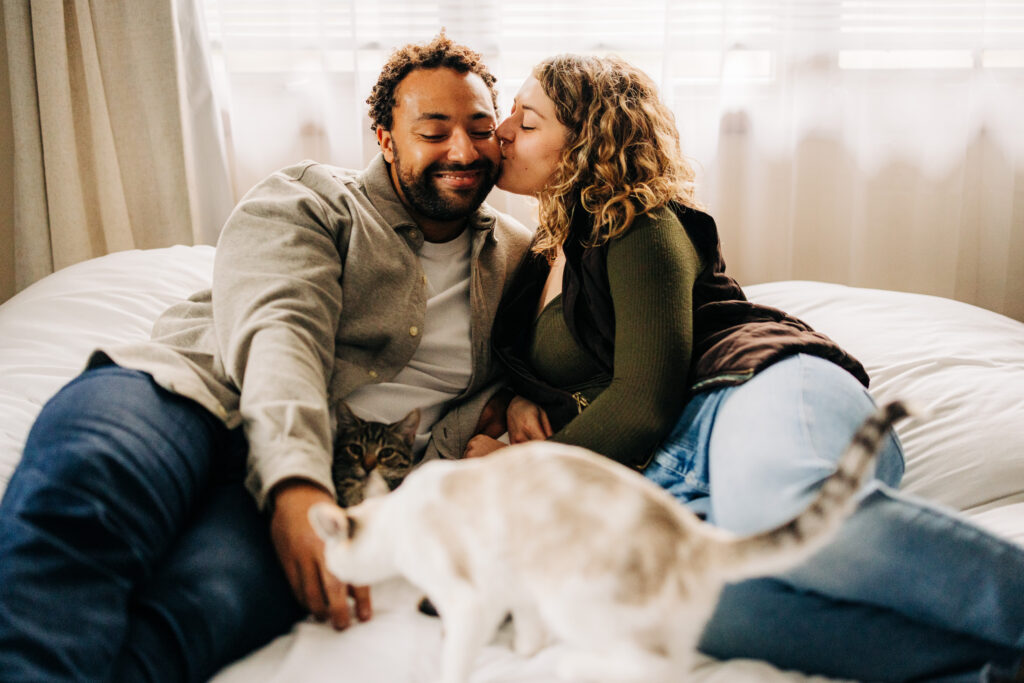 Couple lying on a white bed while one person kisses the other’s cheek, with two cats moving across the bedding.