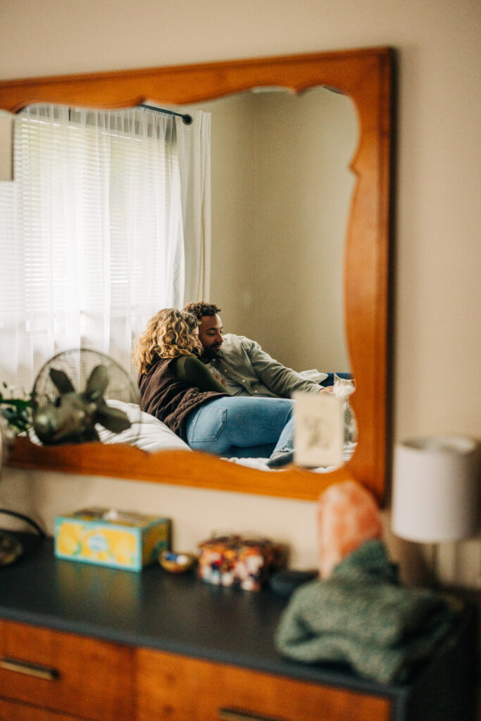 Couple cuddling on a bed seen through a wooden-framed mirror, with soft daylight coming through the window.