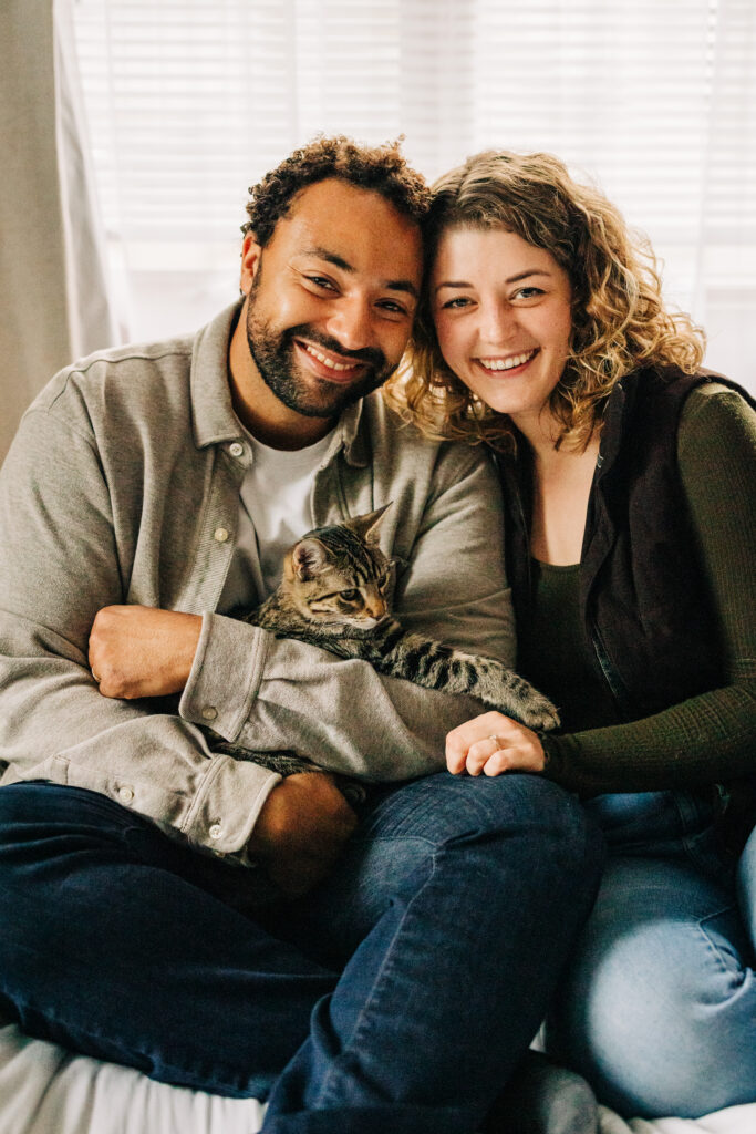 Couple sitting close together on a bed, smiling at the camera while holding a tabby cat between them.