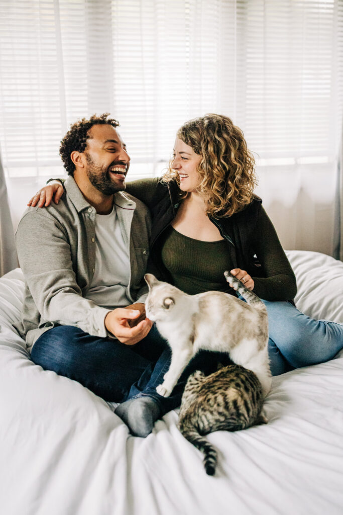 Couple laughing on a bed as two cats walk across the blankets, lit by bright window light.