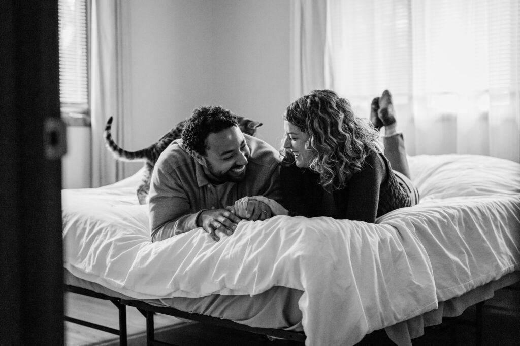 Black-and-white photo of a couple lying on a bed facing each other, smiling, with a cat behind them.