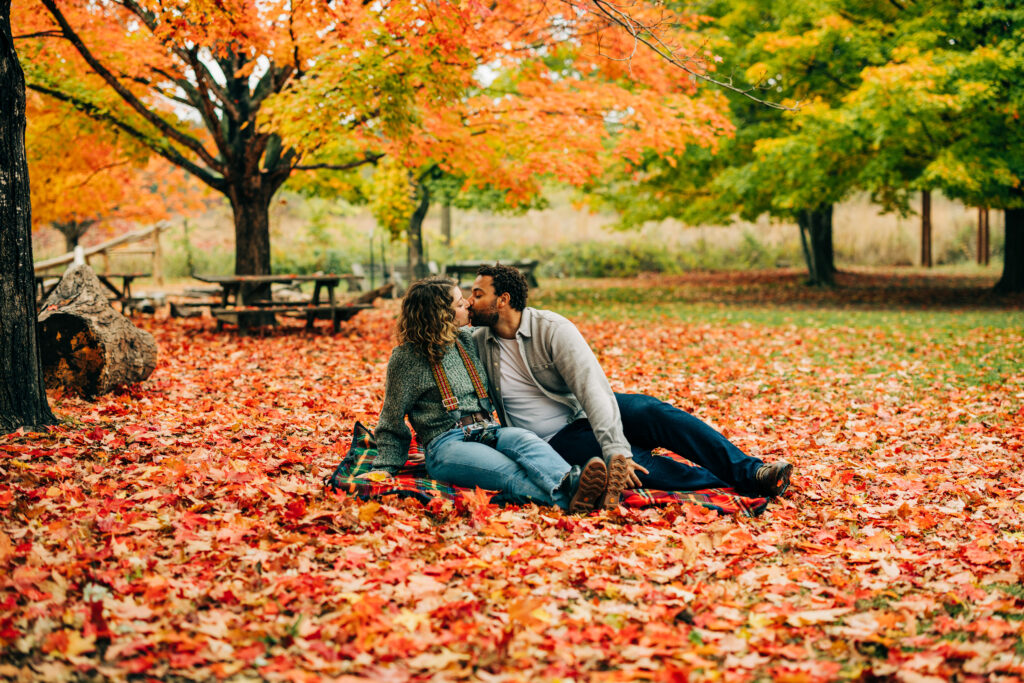 Couple sitting on a blanket in a park covered in fall leaves, sharing a kiss beneath colorful autumn trees.