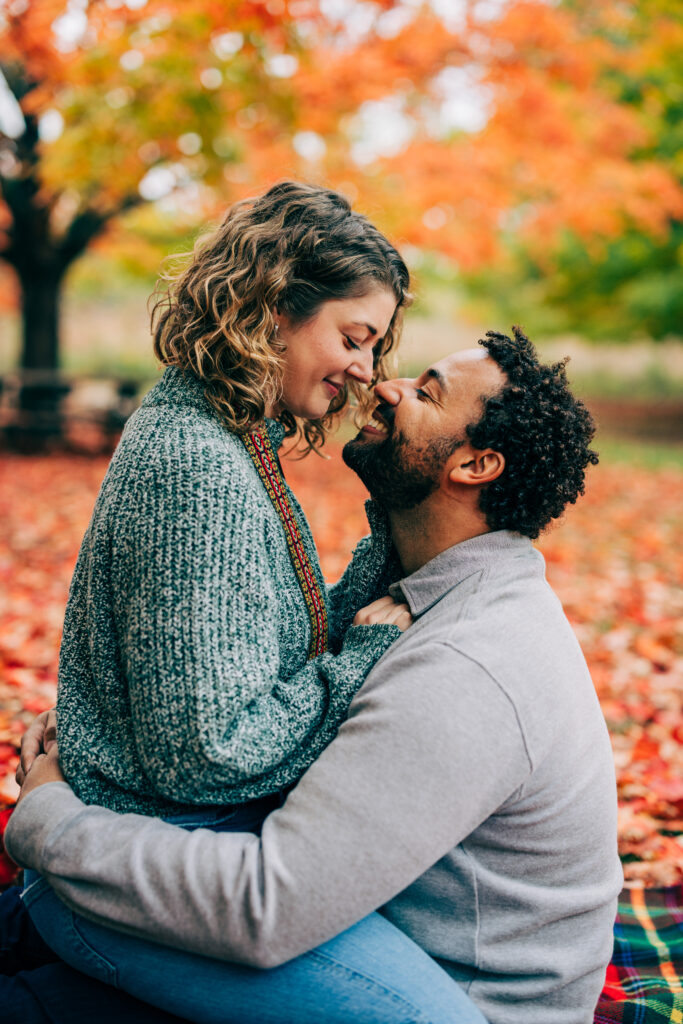 Close-up of a couple embracing outdoors in autumn, surrounded by orange and red leaves in soft natural light.