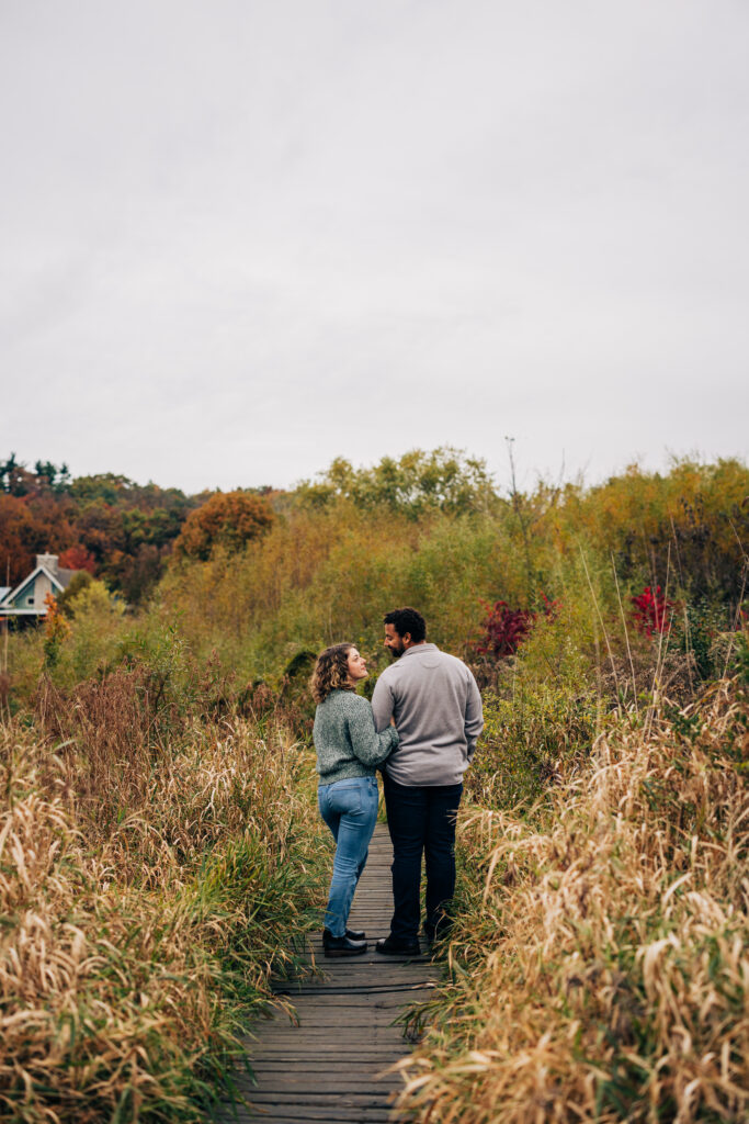 Engaged couple standing on a wooden boardwalk at Aldo Leopold Nature Center, surrounded by prairie grasses and fall color.