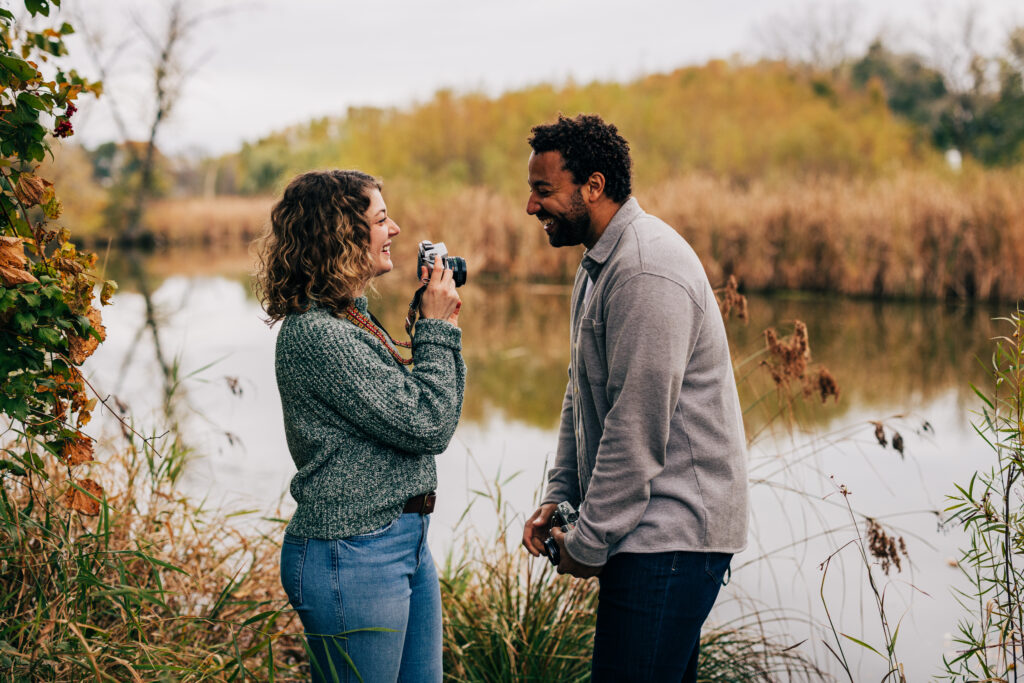Engaged couple smiling at each other beside a pond at Aldo Leopold Nature Center, holding vintage cameras in fall light.