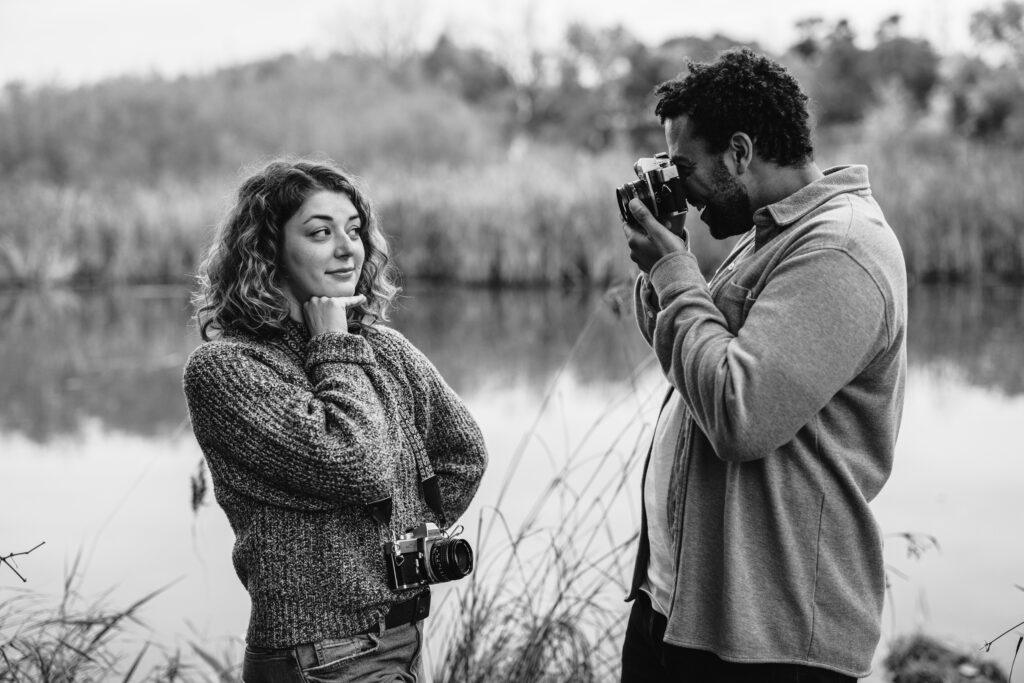 Black-and-white photo of an engaged couple at Aldo Leopold Nature Center, one person photographing the other near water.
