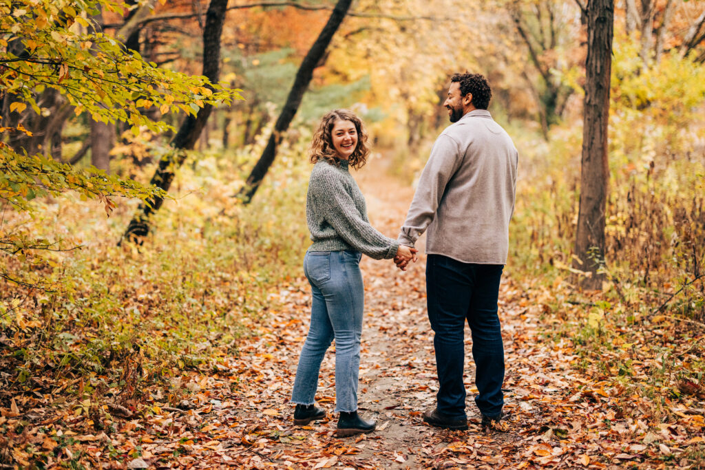 Engaged couple holding hands on a wooded trail at Aldo Leopold Nature Center, surrounded by colorful autumn leaves.