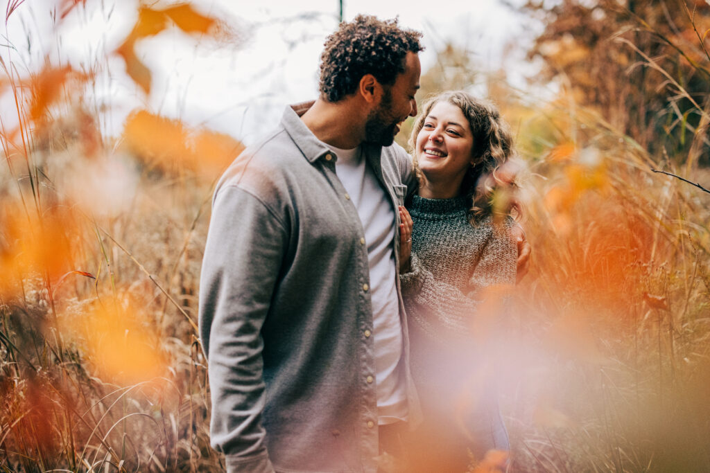 Close-up of an engaged couple standing together in tall grasses at Aldo Leopold Nature Center, framed by soft fall foliage.