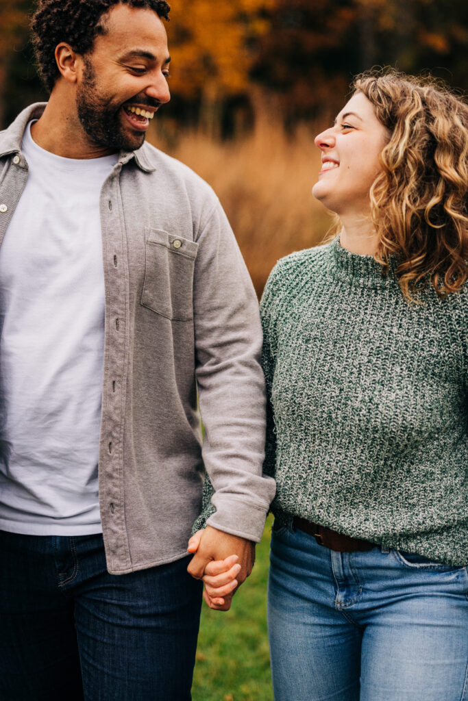 Close-up of an engaged couple holding hands and smiling at each other with fall trees in the background.
